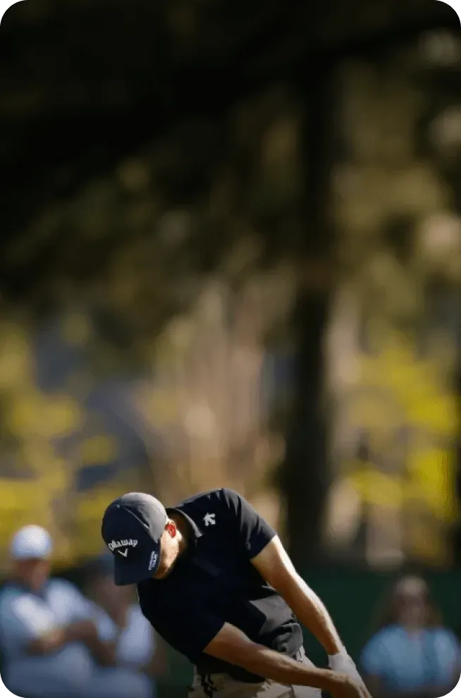 Golfer in black shirt and cap mid-swing with blurred spectators in background.